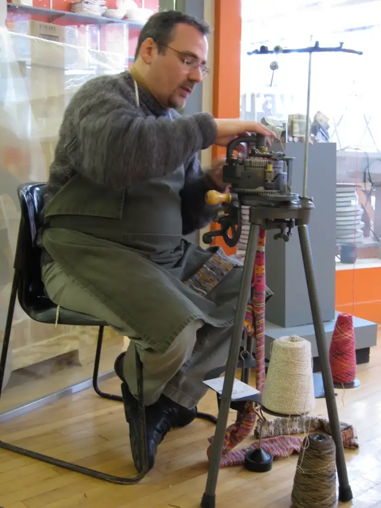 A craftsman knitting a sock on a hand-cranked circular machine, with cones of yarn around him